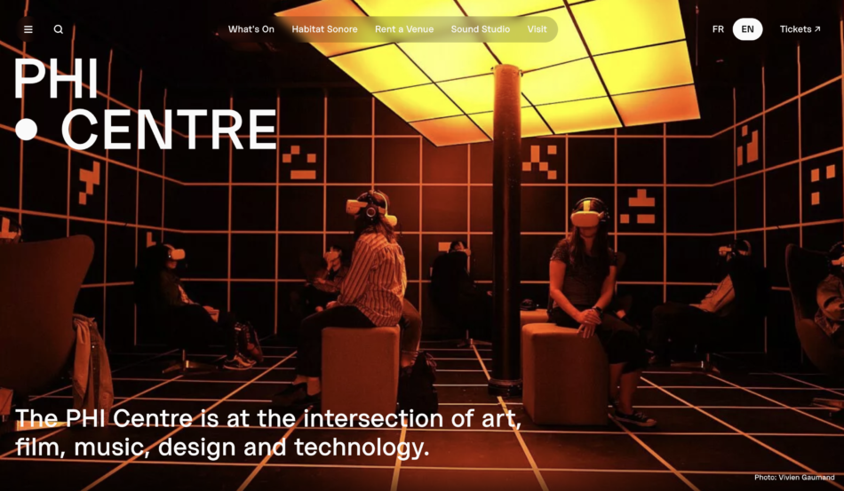 Screenshot of the PHI Centre webpage showing visitors wearing virtual reality headsets in the interactive installation room at the PHI Centre, bathed in an orange light, showcasing the center's focus on combining art, film, music, design, and technology.